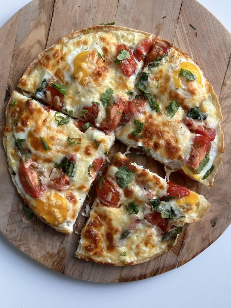 Homemade breakfast pizza with eggs, tomatoes, melted cheese, and herbs on a wooden board.