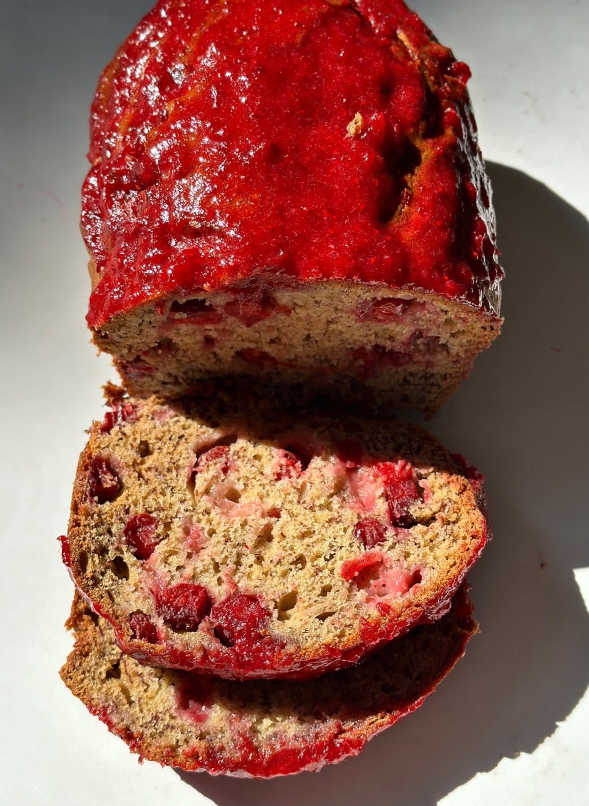 Sliced cranberry banana bread topped with a vibrant cranberry glaze on a white surface.