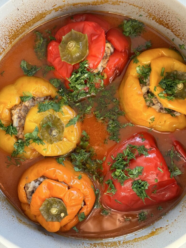 Five large stuffed bell peppers filled with meat and rice, simmering in tomato broth inside a white Dutch oven, garnished with fresh herbs