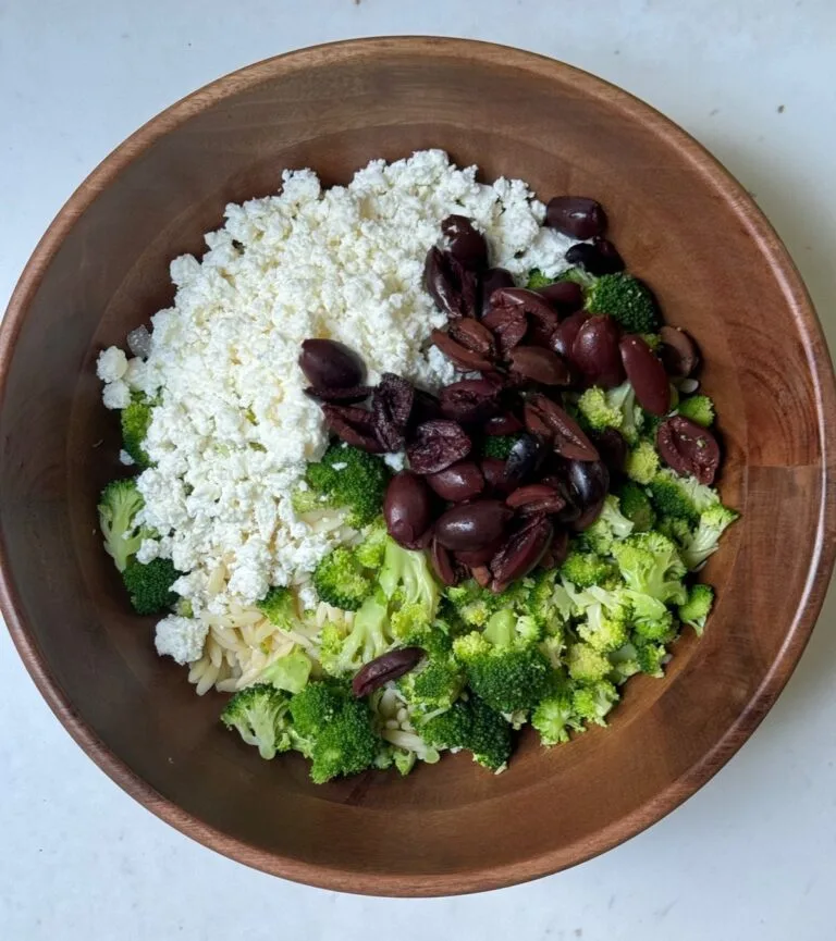 Wooden bowl with orzo broccoli salad topped with feta cheese, broccoli, and Kalamata olives.