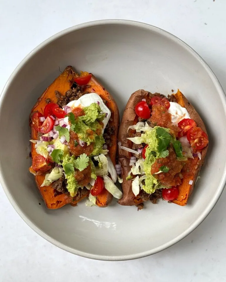 Taco stuffed sweet potatoes topped with ground beef, guacamole, salsa, sour cream, lettuce, tomatoes, and cilantro on a white plate.