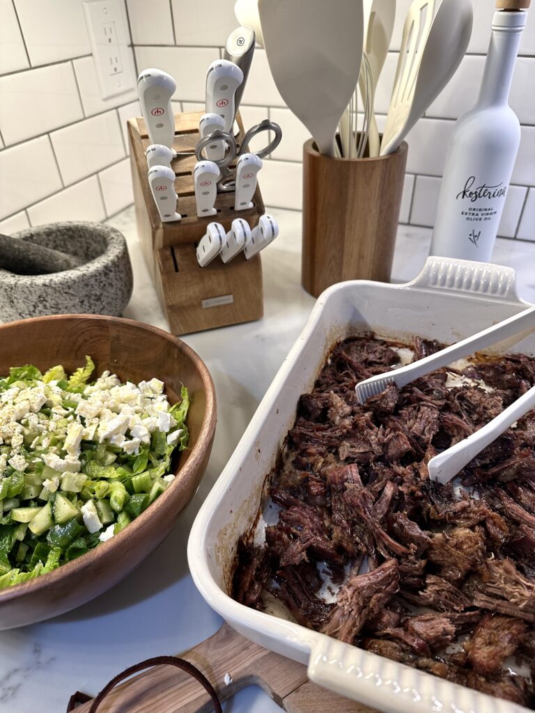 Crispy shredded roast beef in a white baking dish with tongs, next to a wooden bowl of chopped romaine, cucumber, green onion, and feta on a white tile kitchen counter.